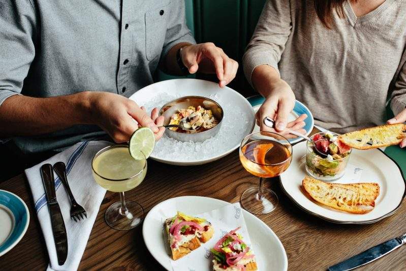 Two diners dig into various dishes including avocado toast and house-smoked salmon dip along with two cocktails at The Darling in Charleston.