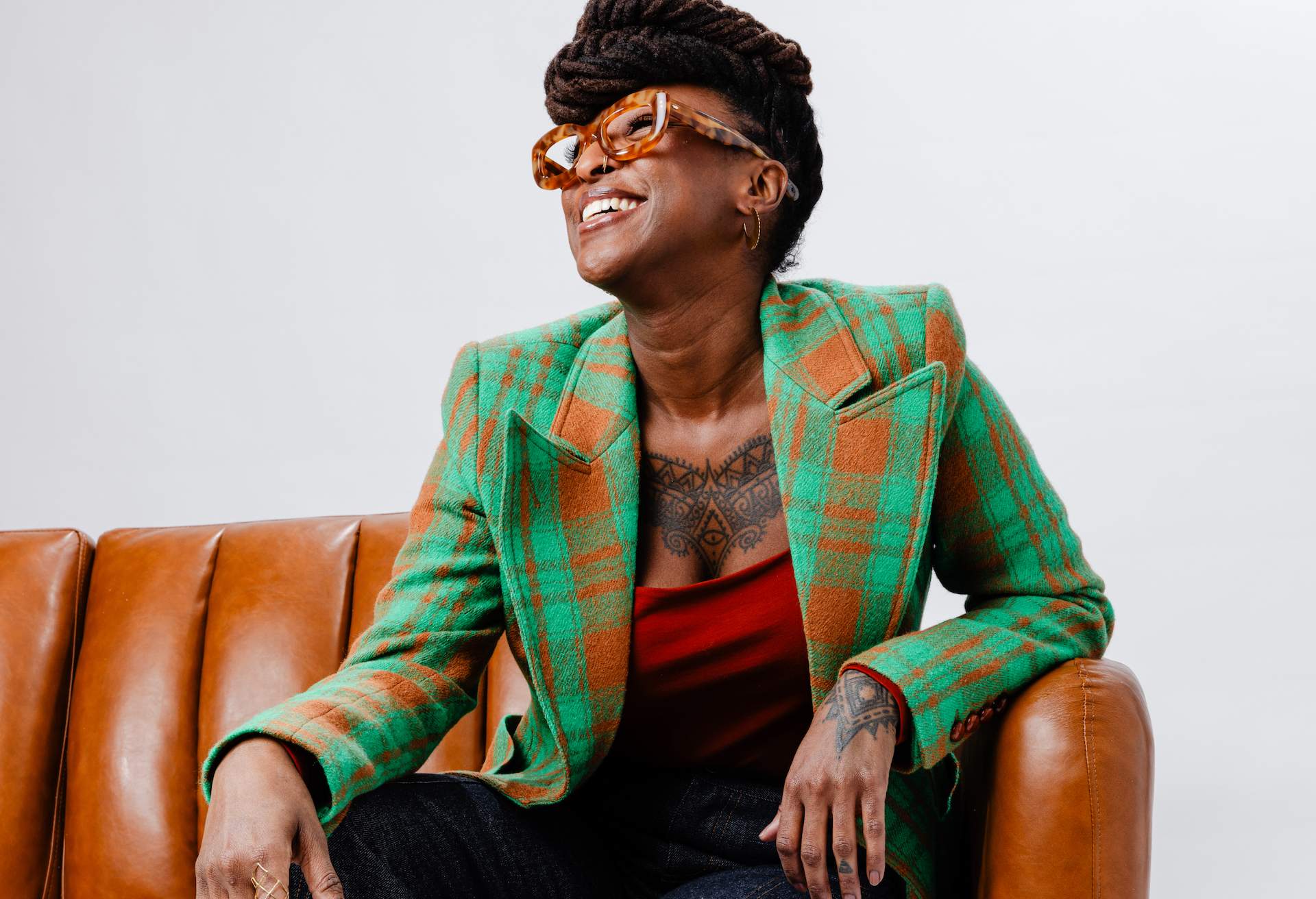 conniesunderground_blogheader_01 Chef Mariya Russell sits on a leather couch wearing a green and orange blazer, looking off to the side and smiling.