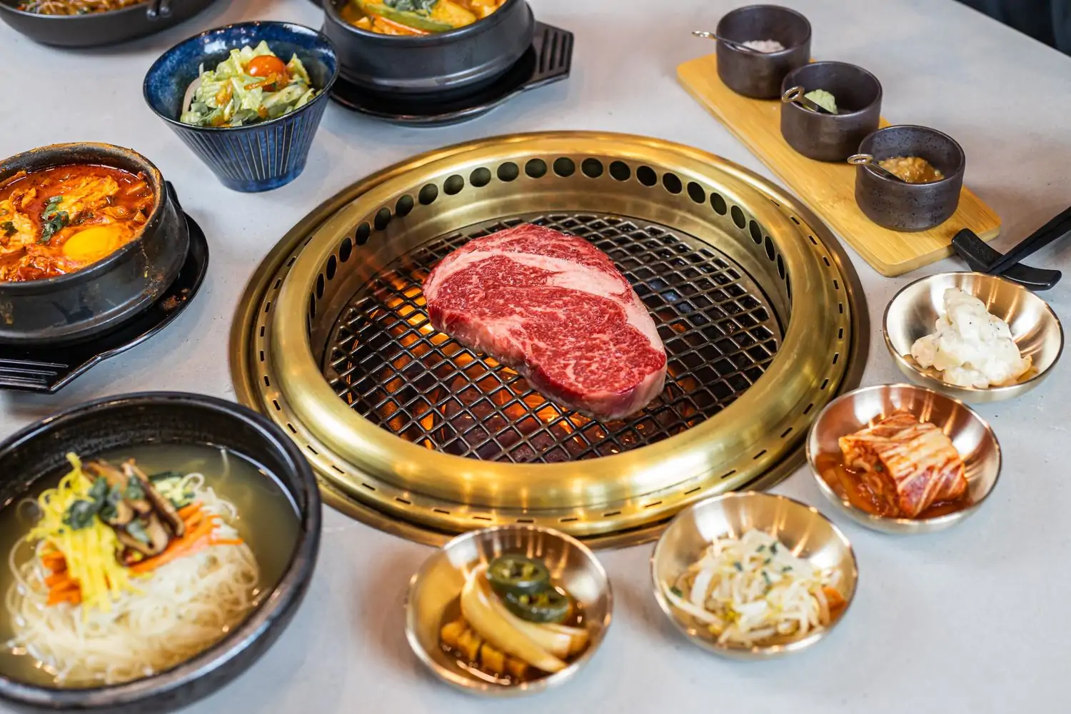 featured image Credit AB Steak by chef Akira Back.jpeg A table setting with a grill in the center with a piece of meat cooking over the flame surrounded by side dishes like salad and noodles and sauces at AB Steak by chef Akira Back, one of the best Korean restaurants in Los Angeles found on OpenTable.