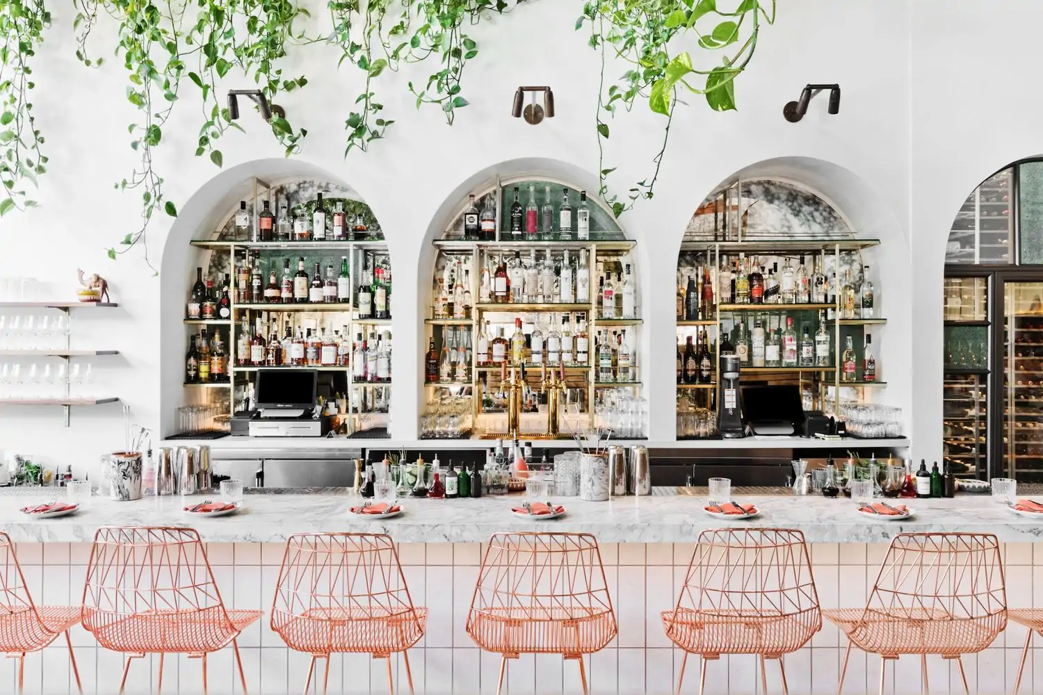 Credit Bavel.jpg A stylish restaurant bar with a white marble countertop, rose gold barstools, and a wall lined with shelves displaying an array of liquor bottles. The bar is decorated with lush greenery and arched windows at Bavel, one of the best Middle Eastern restaurants in Los Angeles on OpenTable.