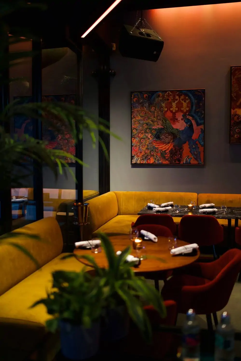 Credit Basbussa.jpg A dimly lit restaurant interior with a vibrant peacock painting on the wall, a yellow velvet banquette, and a round table with red chairs at Basbussa in West Hollywood, Los Angeles, available to reserve on OpenTable.