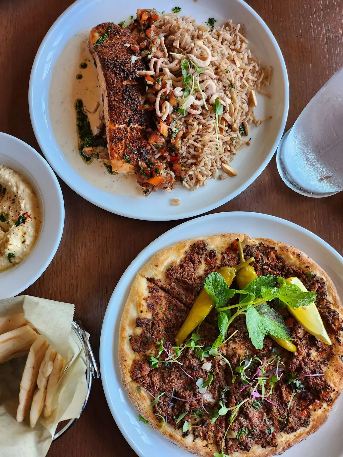 Credit Levant Bistro + Bake Shop.jpeg A top-down view of a table with two plates of Middle Eastern food. One plate has a piece of grilled salmon with rice and vegetables; the other has a flatbread topped with za'atar and garnished with peppers and mint. There is also a bowl of hummus and a basket of bread on the table at Levant Bistro + Bake Shop in Los Angeles, available to reserve on OpenTable.