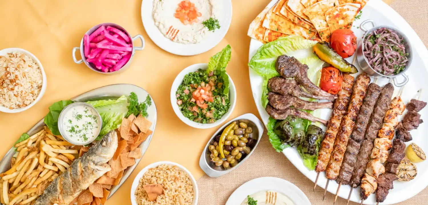 Credit Carousel Restaurant.png A top-down view of a table spread with a variety of Middle Eastern dishes, including grilled meats, rice, salads, dips, and bread atop a light wood table at Carousel Restaurant, one of the best Lebanese restaurants Los Angeles has on OpenTable.