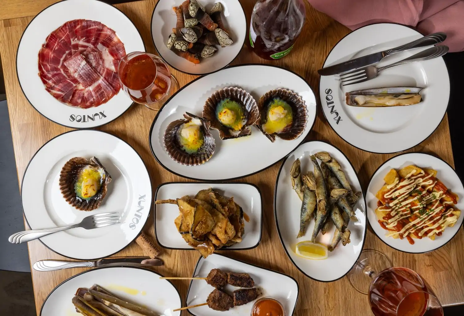 A wood table filled with a variety of Spanish tapas, including cured ham, grilled seafood, fried fish, and potato dishes. There are also glasses of wine on the table at Xuntos in Santa Monica, available to reserve on OpenTable.