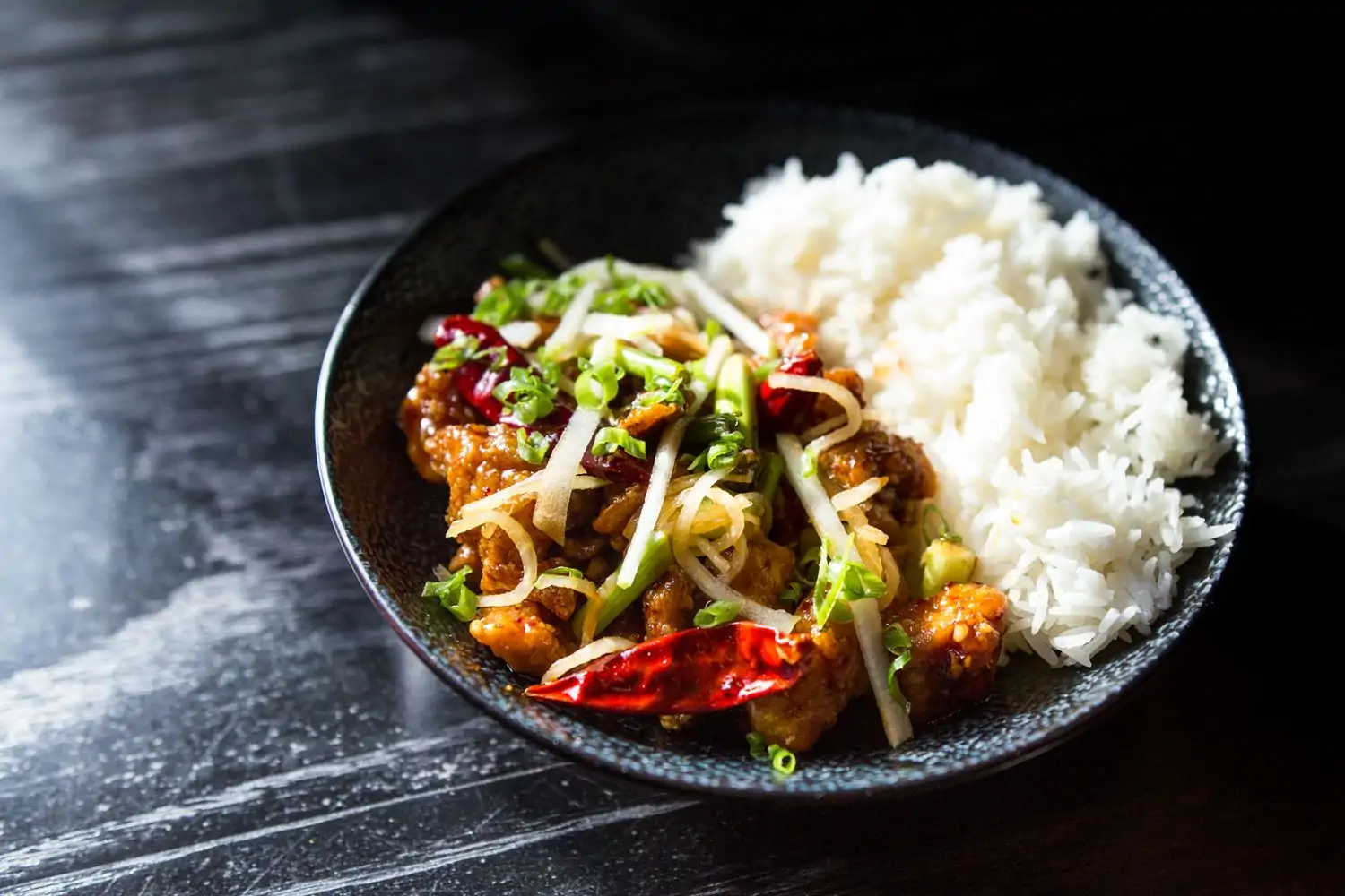 A black dish of kung pao chicken with sliced green onions and red chili peppers, served with steamed rice at Old Thousand, one of the best Asian restaurants in Austin, Texas, available on OpenTable.