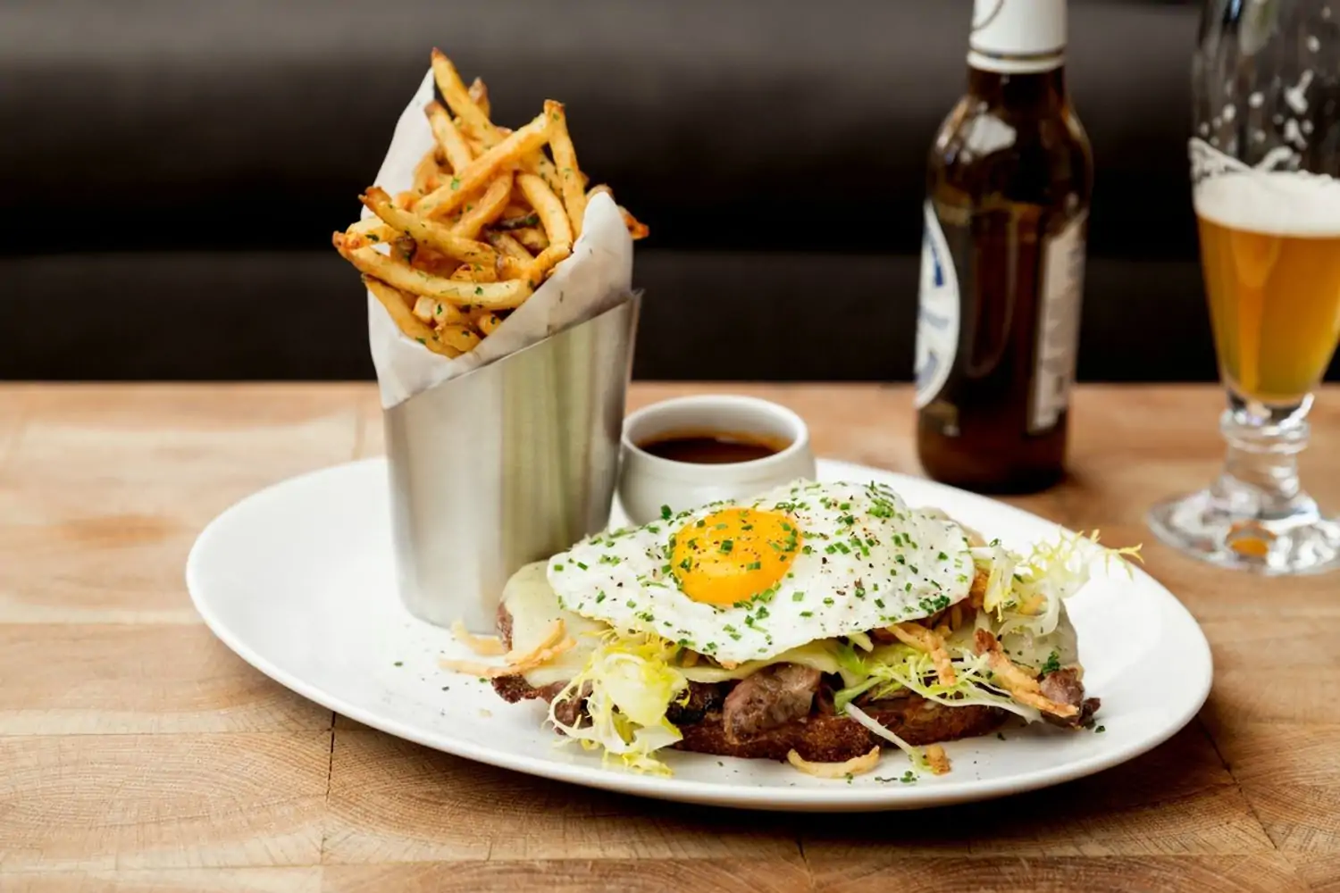 Croque Madame with a fried egg, fries in a metal cup, and a beer on a wooden table at Brentwood’s Baltaire, the best boozy brunch Los Angeles on OpenTable.
