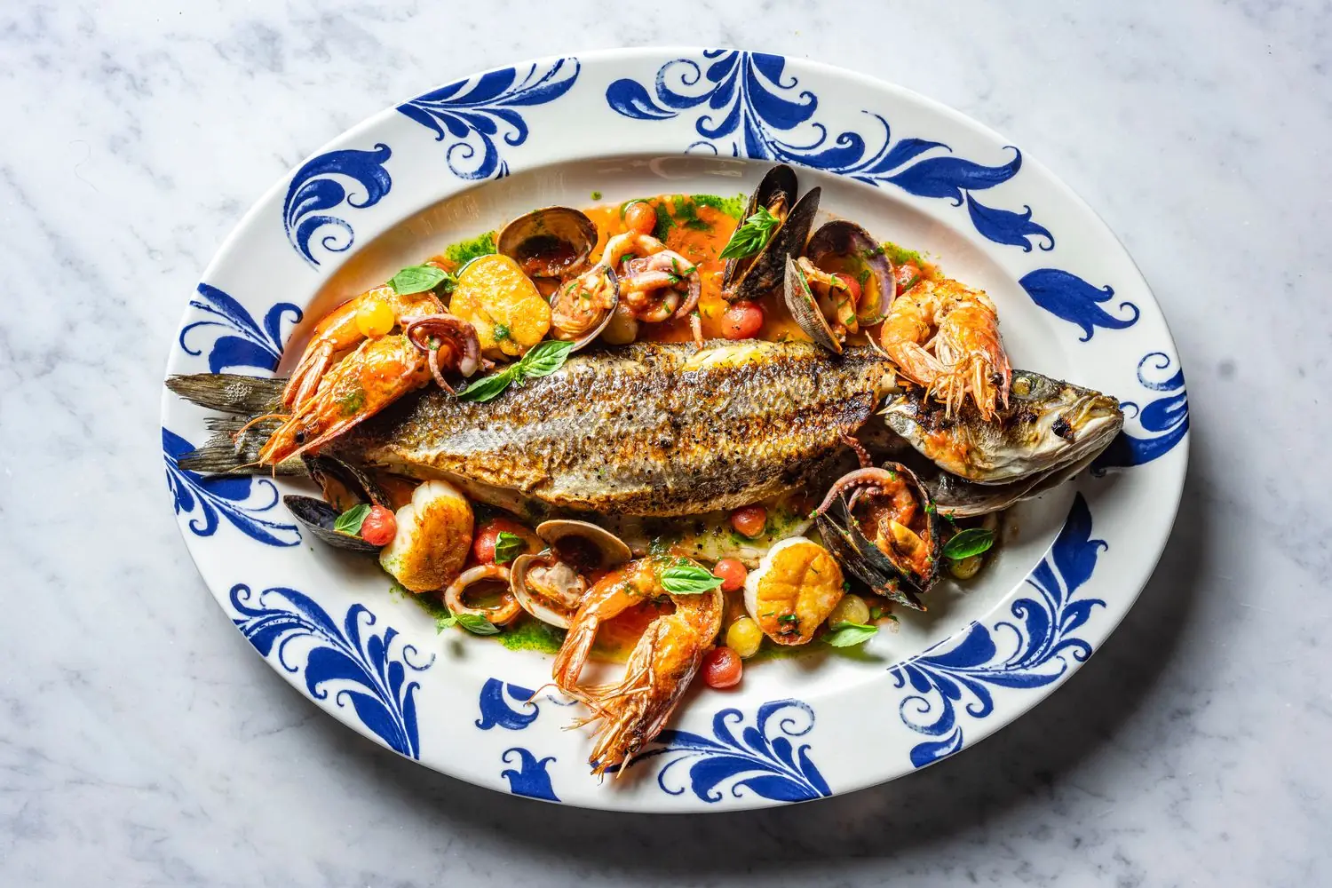A whole roasted fish with scallops and other seafood around it on a blue plate at Tre Dita in Chicago