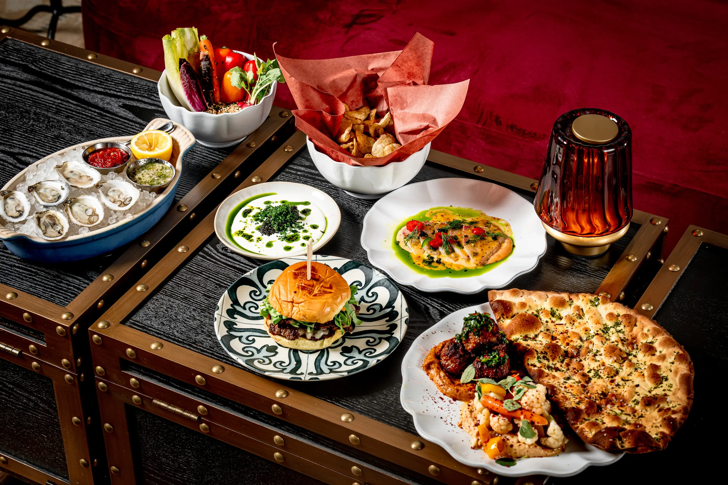 A full table spread at Golden Child in NYC featuring the Golden Burger, Montauk Pearl oysters, hamachi, roasted meatballs with naan, seasonal crudité, caviar, and potato chips, arranged on a brass-trimmed dark wood tray.