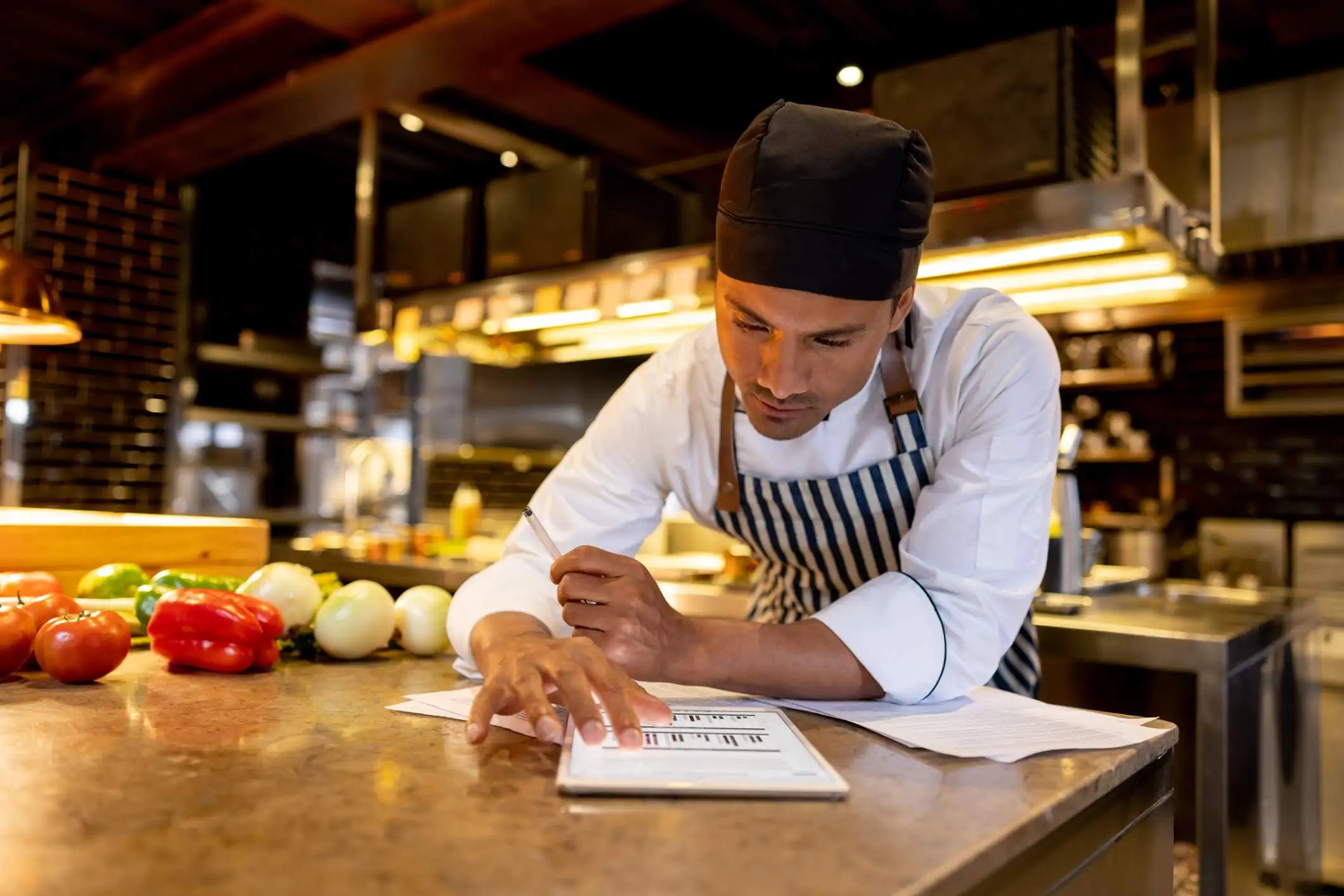 2022-crm-sup2 Image depicts a person wearing a black and white striped apron and black cap leaning over a countertop. They are examining graphs on a tablet and there are sheets of paper splayed under them. In the background is an industrial kitchen. There are vegetables sitting on the counter next to them.
