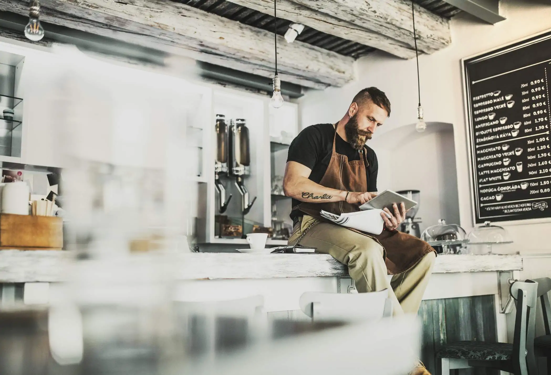 2022-crm-sup1 Image depicts a person sitting on a countertop using a tablet. Next to them sits a cup of coffee and a notebook. There is a coffee menu in the back. The establishment is very industrial, with light bulbs hanging from the ceiling.