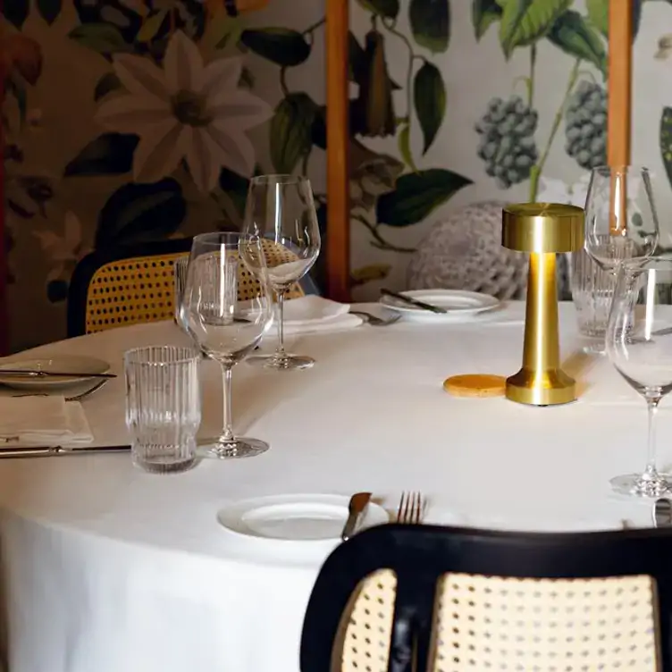 le-crocodile4webp A round, white-clothed table set with cutlery, side plates and glasses, and surrounded by floral wallpaper at Le Crocodile.